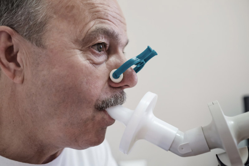 Immagine di un uomo anziano con baffi con il naso tappato da una speciale molletta (stringinaso) e con in bocca un boccaglio, attaccato a spirometro, mentre svolte una pletismografia