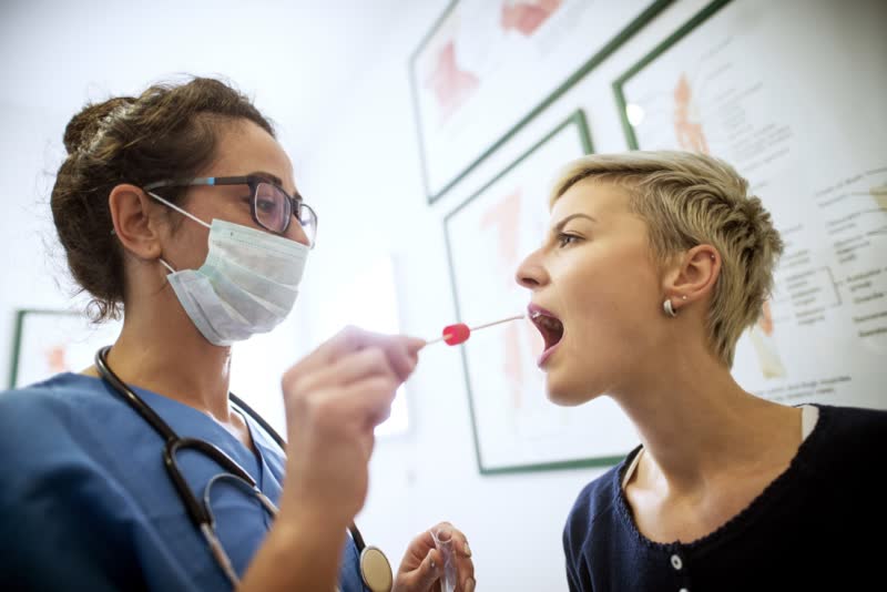 Infermiera in camice azzurro con mascherina mentre con un tampone preleva un campione di saliva da una giovane paziente bionda per effettuare un test del dna 