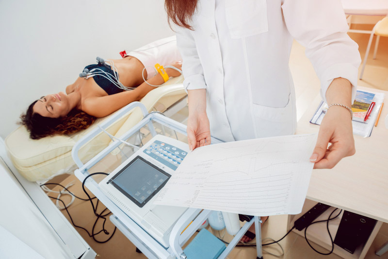 Paziente donna sdraiata su un lettino di una clinica mentre esegue un elettrocardiogramma che una dottoressa sta leggengdo 