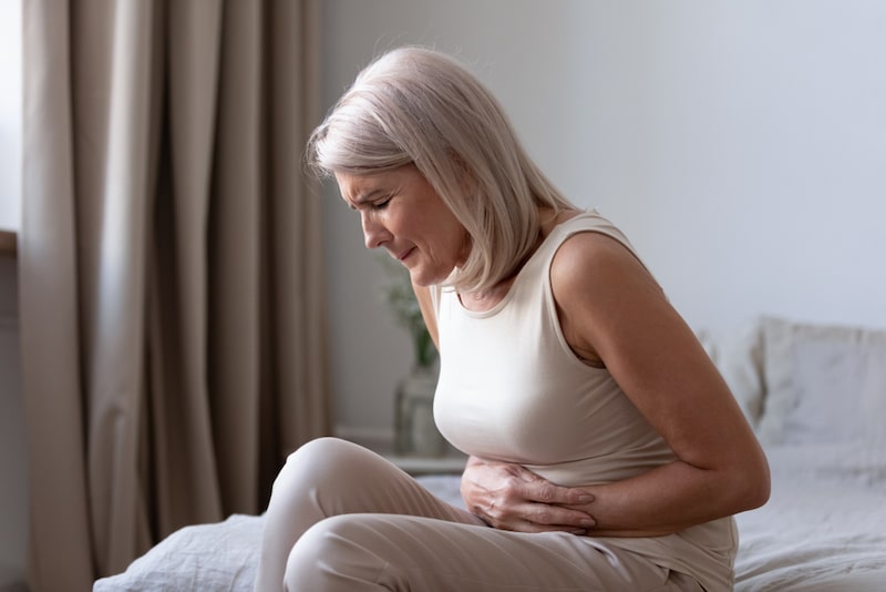 Foto di una donna seduta su un letto mentre si stringe la pancia per i dolori da blocco intestinale