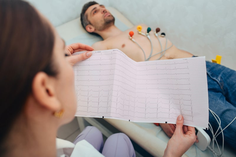 Una dottoressa tiene in mano i risultati dell'ECG e sullo sfondo si vede il paziente sdraiato su un lettino 