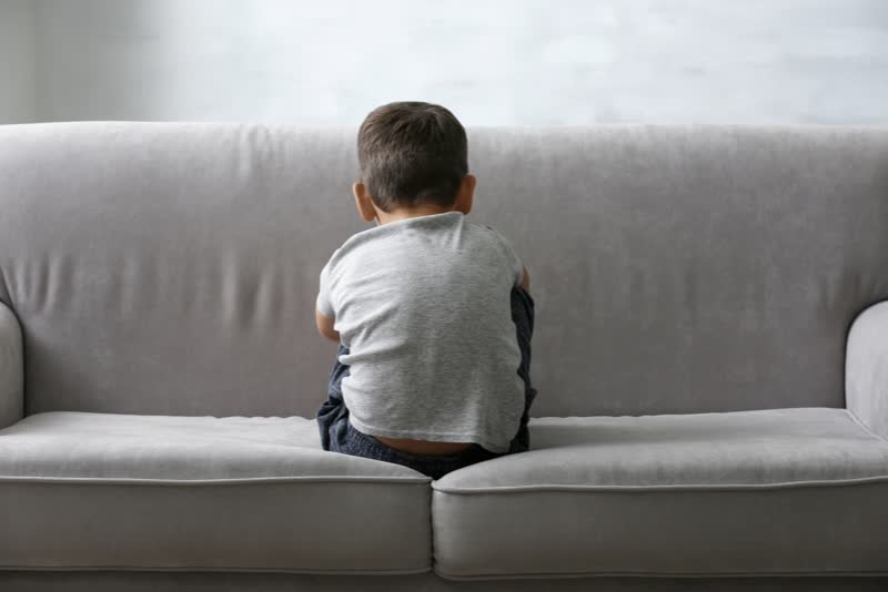 Foto di bambino affetto da disturbi dello spettro autistico (autismo) seduto su un divano grigio e vestito con maglietta di colore chiaro di spalle che dà le spalle alla fotocamera mentre pensa