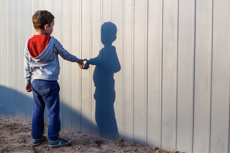 Foto di bambino affetto da autismo in piedi, vestito con jeans e felpa grigia che guarda verso una staccionata mentre gioca con la sua ombra con la sua mano destra
