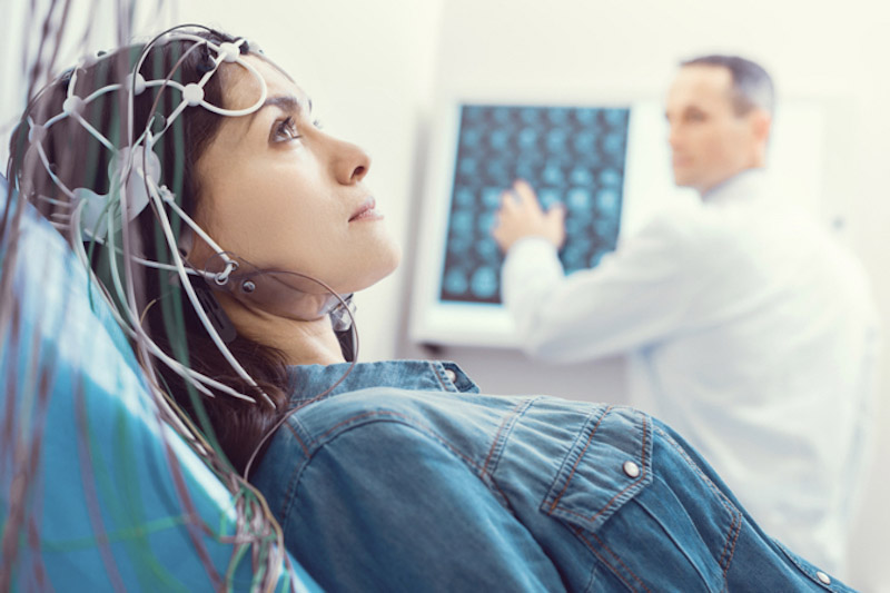 Primo piano di una donna in una sala di ospedale con elettrodi in testa per effettuara un'elettroencefalografia mentre il medico sullo sfondo attiva il macchinario