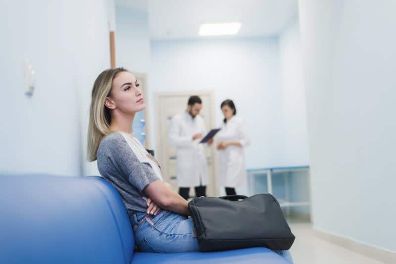 Donna giovane bionda seduta con al suo fianco una borsa nera nella sala di attesa di una struttura ospedaliera (ospedale o ambulatorio) in attesa di effettuare una cistografia