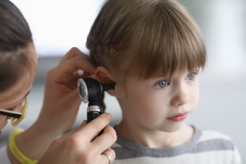 Foto di dottore otorino/otorinolaringoiatra in primo piano con otoscopio utilizzato per osservare all'interno dell'orecchio di una bambina con gli occhi azzurri e i capelli a caschetto castani per vedere la presenza di otite