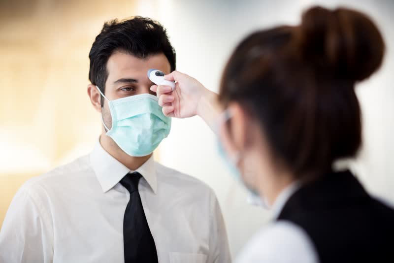 Foto di donna che misura la temperatura corporea ad un uomo con mascherina azzurra per vedere se ha la febbre