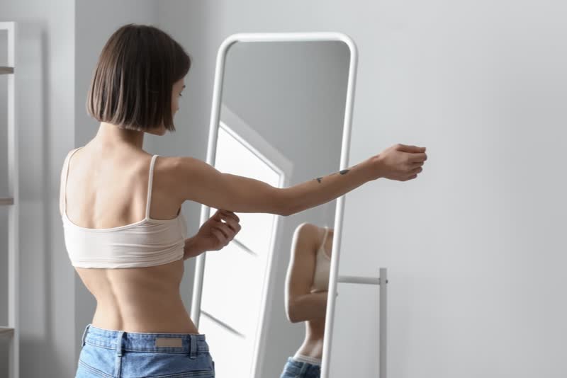 Foto di donna magra di spalle con reggiseno mentre si guarda allo specchio e si pizzica il tricipite del braccio destro per osservare il livello di grasso, per simboleggiare le difficoltà dell'anoressia nervosa