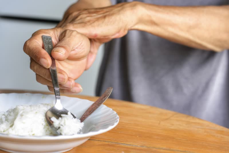 Foto di persona anziana intenta a mangiare del riso che deve tenersi con la sinistra la sua mano destra mentre cerca di raccogliere del riso dal piatto col cucchiaio a causa del morbo di parkinson di cui soffre