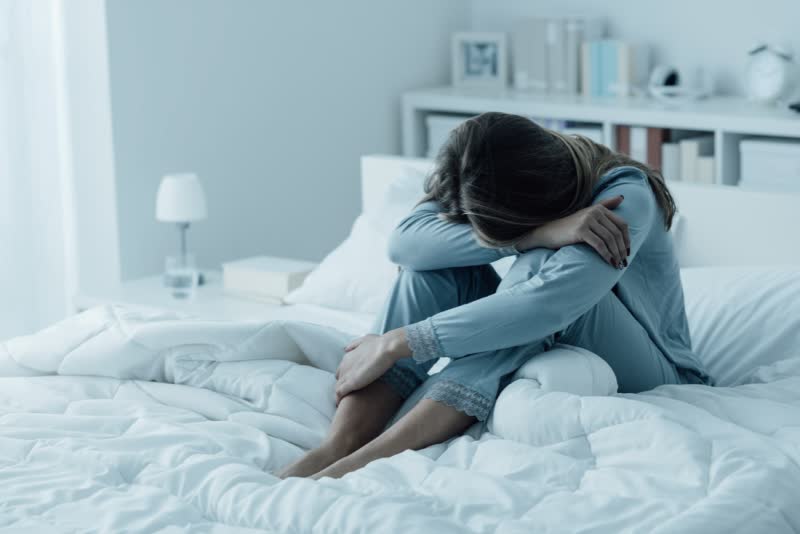 Foto di donna seduta su un letto con le braccia appoggiate sulle ginocchia e che coprono il viso perché affetta da depressione