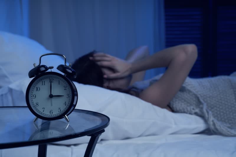 Foto scura di donna con le mani sul volto perché non riesce a dormire per colpa dell'insonnia mentre in primo piano si vede una sveglia che segna le tre 3 di notte