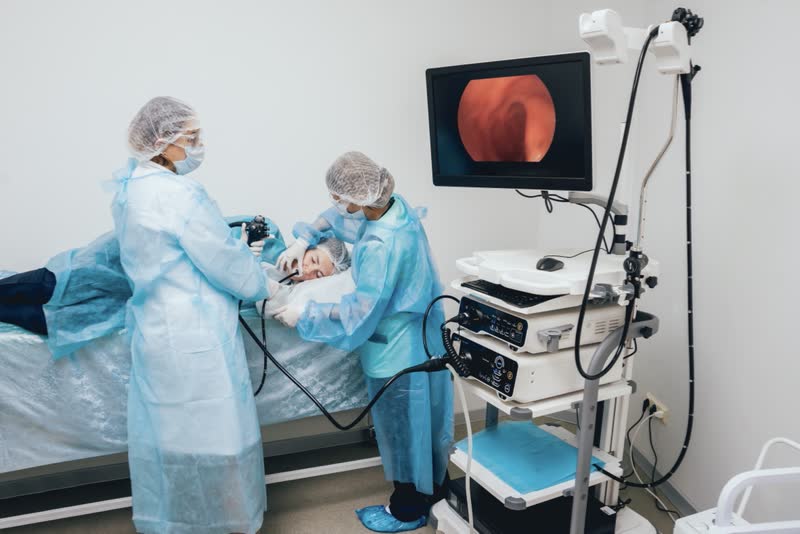 Equipe medica che effettua una gastroscopia su paziente disteso su un lato. Su monitor si vede l'imaging del test