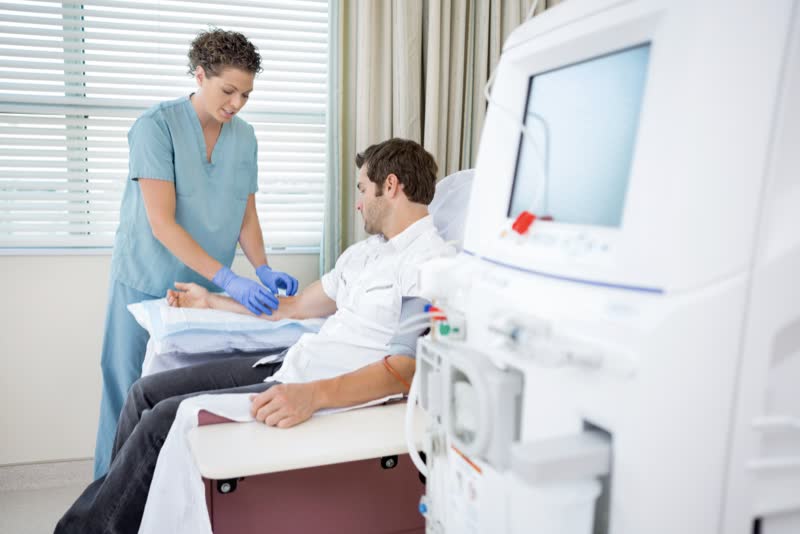 Macchinario per la dialisi in primo piano, in secondo piano un'infermiera in camice azzurro applica la dialisi su un paziente seduto vestito di bianco