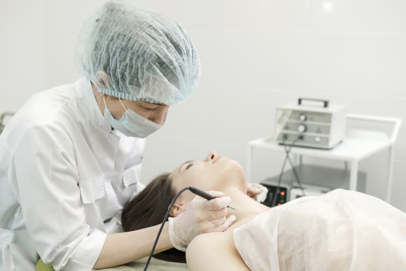 Terapeuta in camice bianco, mascherina e cuffia per i capelli che applica la diatermocoagulazione su una paziente giovane vestita di bianco sdraiata su un lettino di una sala ambulatoriale o ospedaliera